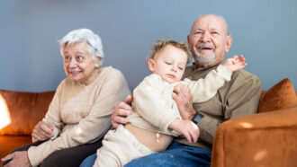 Two elderly people, who are more at risk of dementia, sit on a rust-colored couch. One of them is holding a small child.